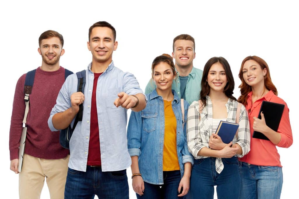 A diverse group of happy adult learners, with one male student in the foreground pointing directly at the camera, holding books, backpacks, and a tablet against a white background.